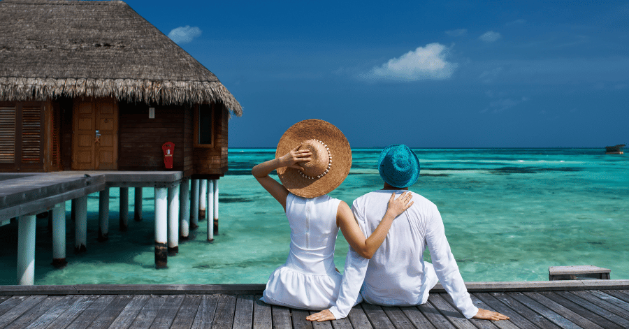 A couple in white sitting on a wooden jetty beside a thatched overwater villa, looking over a turquoise lagoon