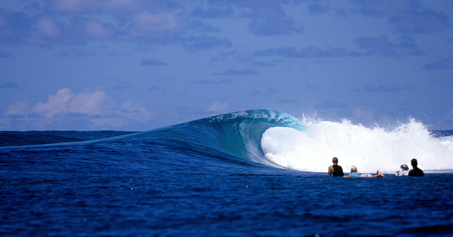 A blue wave breaking over a reef with a few surfers waiting in the lineup