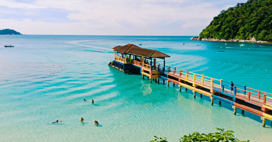 Colourful wooden jetty over clear turquoise water with swimmers, framed by forested headlands in the Perhentians