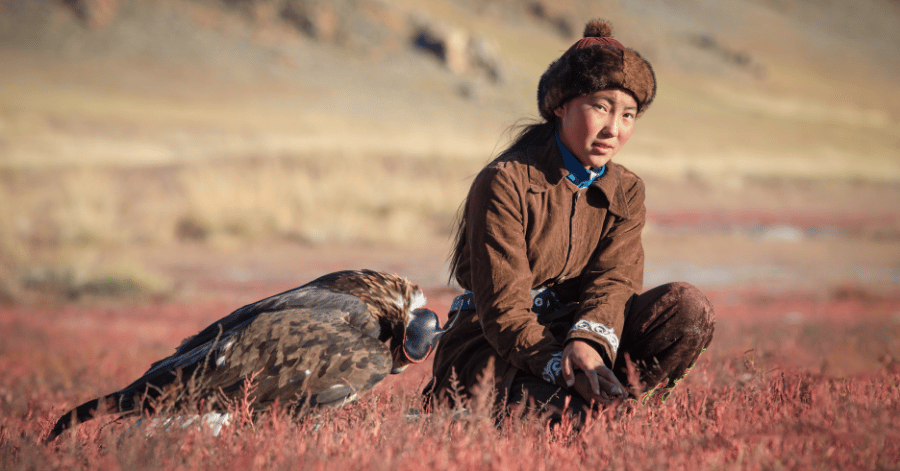 A young Kazakh eagle hunter kneeling beside a golden eagle on the steppe