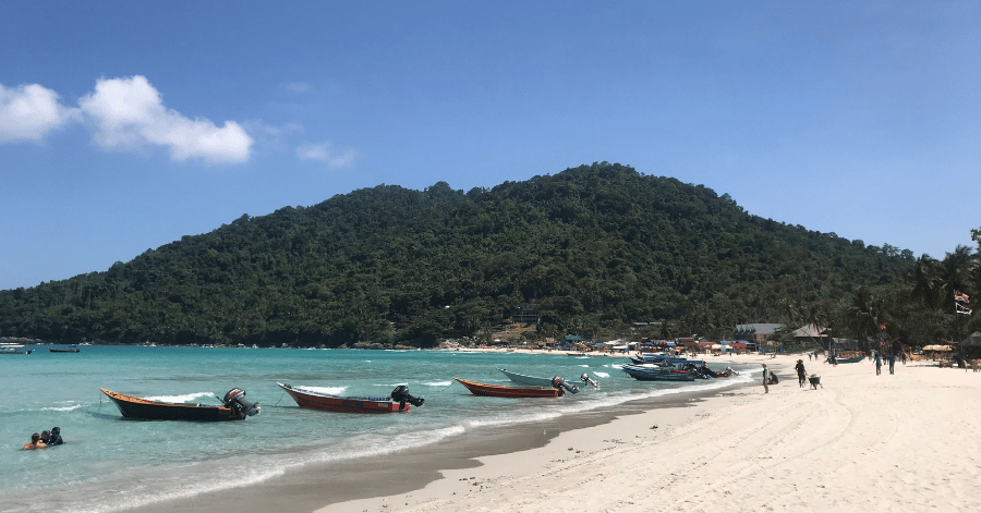 Small boats moored along a white-sand Perhentian beach below a forested hill on a clear, sunny day