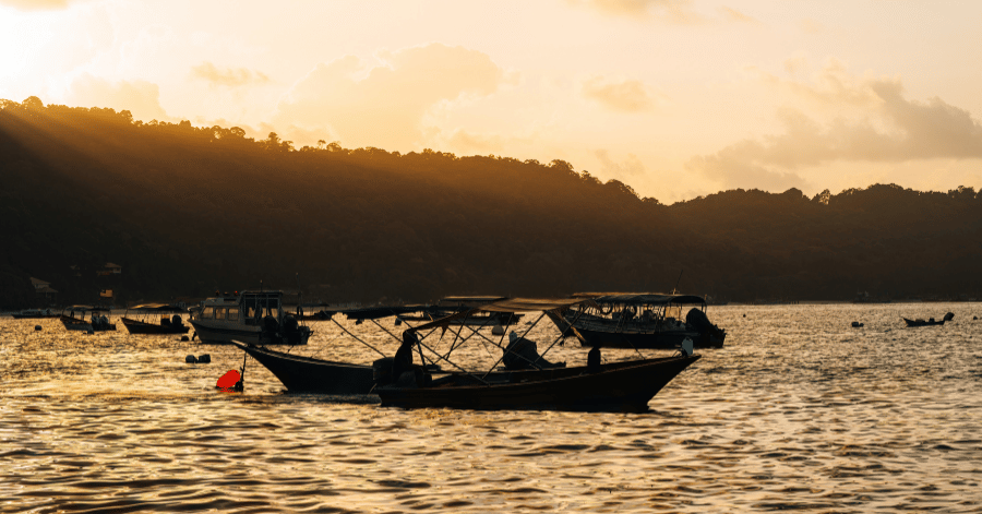 Silhouetted boats moored on calm water at golden hour, with forested hills behind the Perhentian Islands