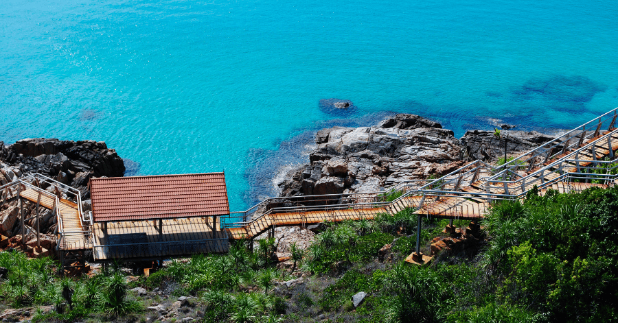 A wooden coastal boardwalk and red-roofed pavilion set on rocks above bright turquoise water