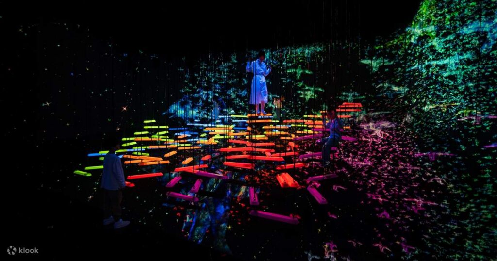 Visitors in a dark room with projected flowers and arcing blue light at teamLab Planets