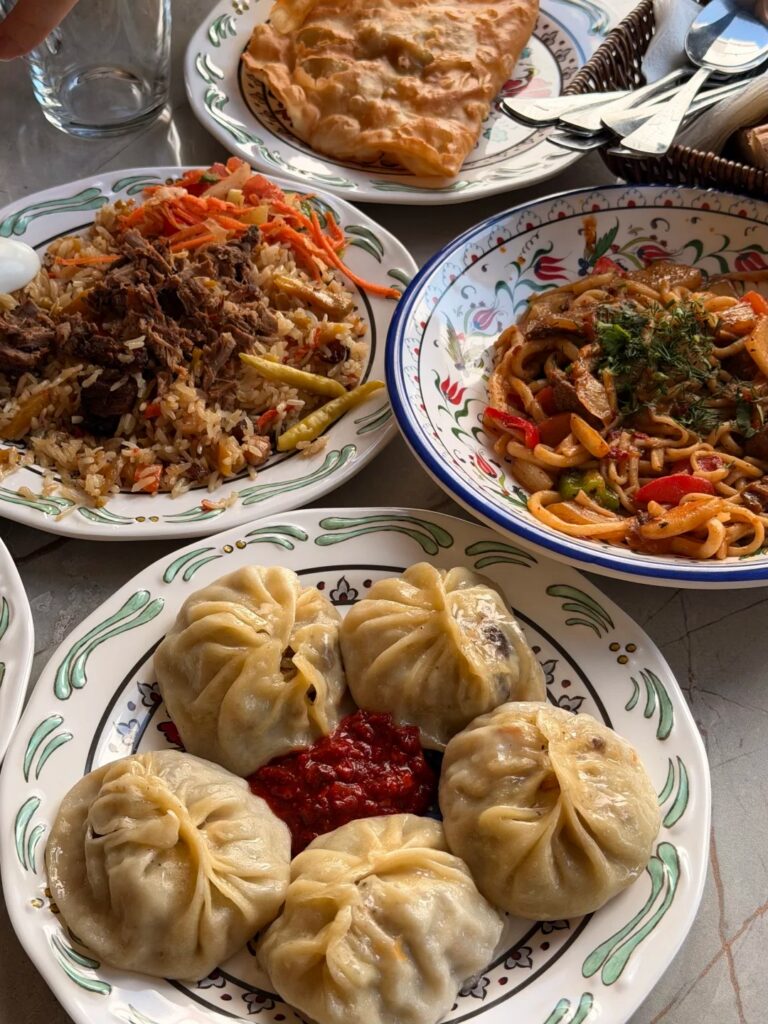 A plate of steamed manti dumplings with red sauce beside plov and noodles