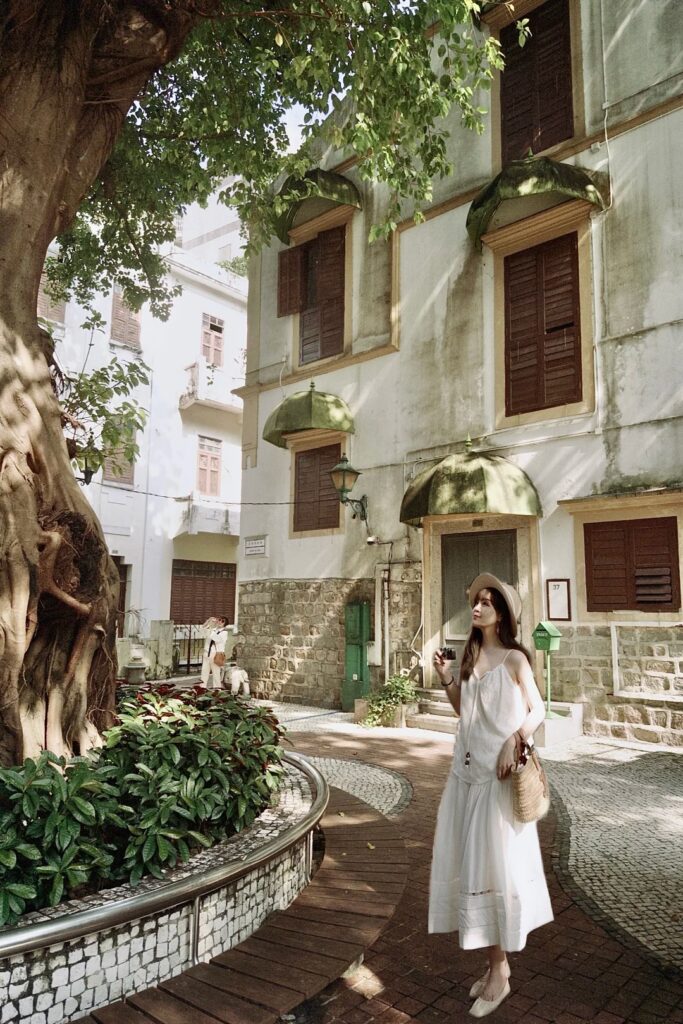 A woman in a white dress strolls a quiet Macau Peninsula lane lined with shuttered colonial buildings