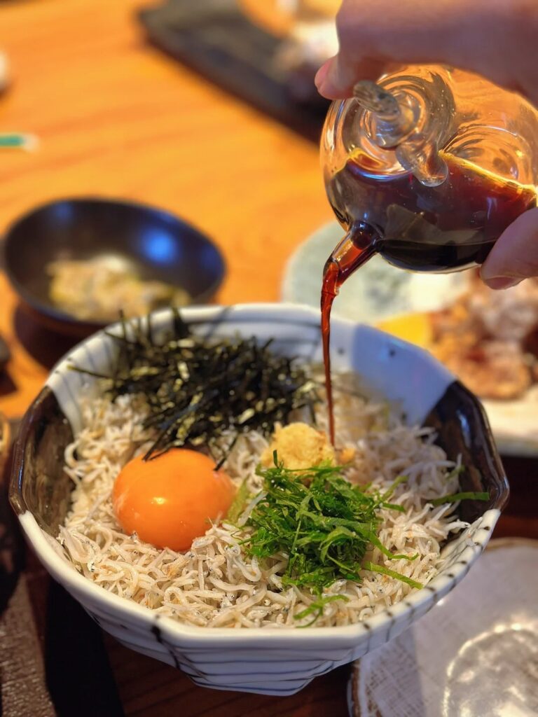 A Kamakura shirasu rice bowl with whitebait, nori and a raw egg yolk, soy sauce being poured over