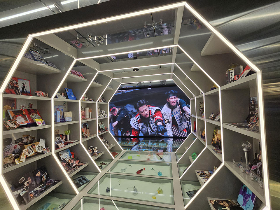Hexagonal white shelving displaying K-pop albums and merchandise around a video screen