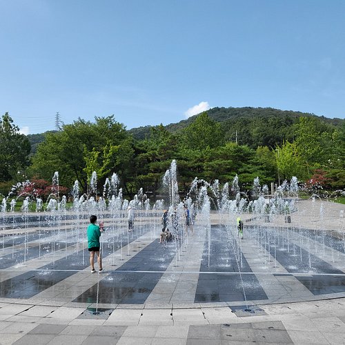 Children running through jets of water on a chequered stone plaza with wooded hills behind