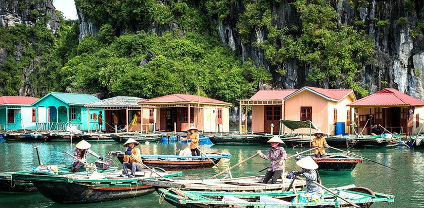 Women in conical hats rowing wooden boats past pastel floating houses below a cliff