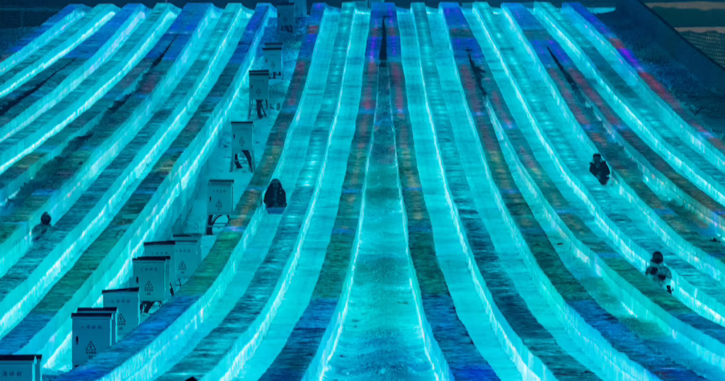 Long illuminated blue ice slides at night with people sliding down the chutes