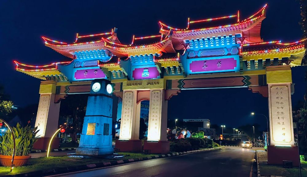 Illuminated Chinese ceremonial arch with clock tower spanning Jalan Padungan at night