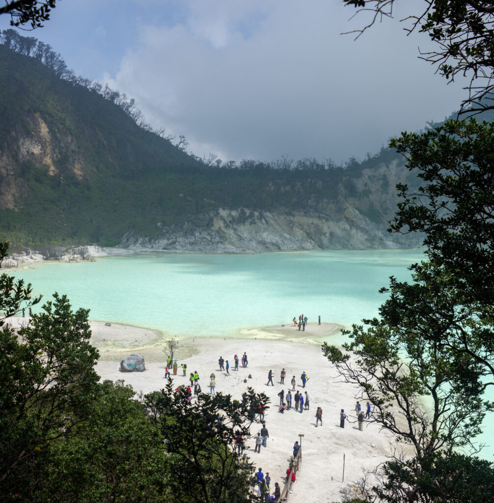 Visitors on the white mineral shore of Kawah Putih's turquoise crater lake, framed by trees