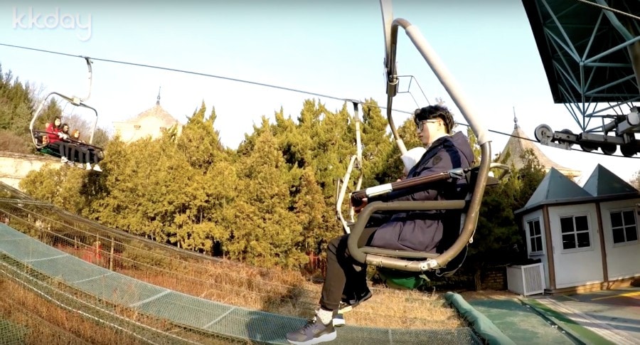 A visitor rides an open chairlift above safety netting and conifers at Everland