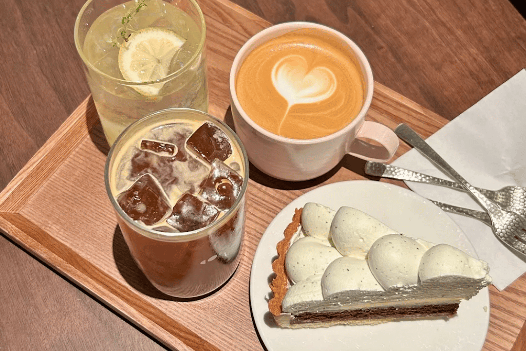 Latte with heart art, iced coffee and lemonade on a wooden tray beside a cream tart