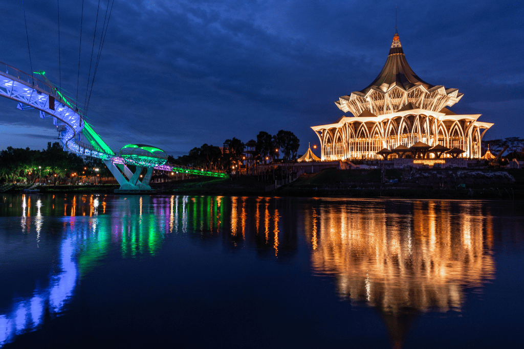 Darul Hana Bridge lit blue and green beside the floodlit State Assembly across the Sarawak River