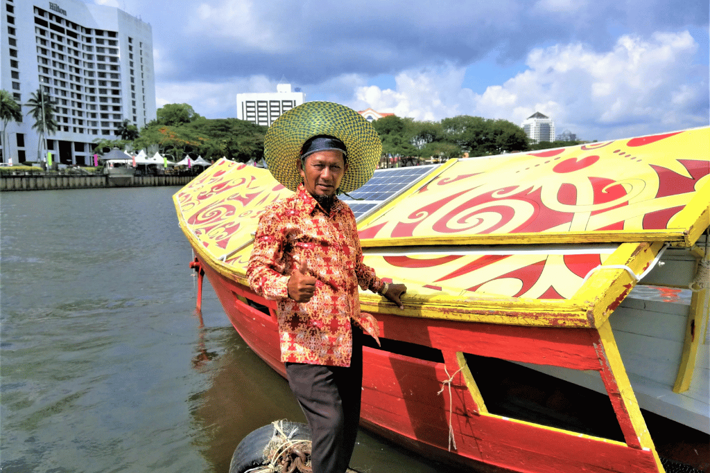Boatman in batik shirt and woven hat beside a red and yellow painted tambang river boat