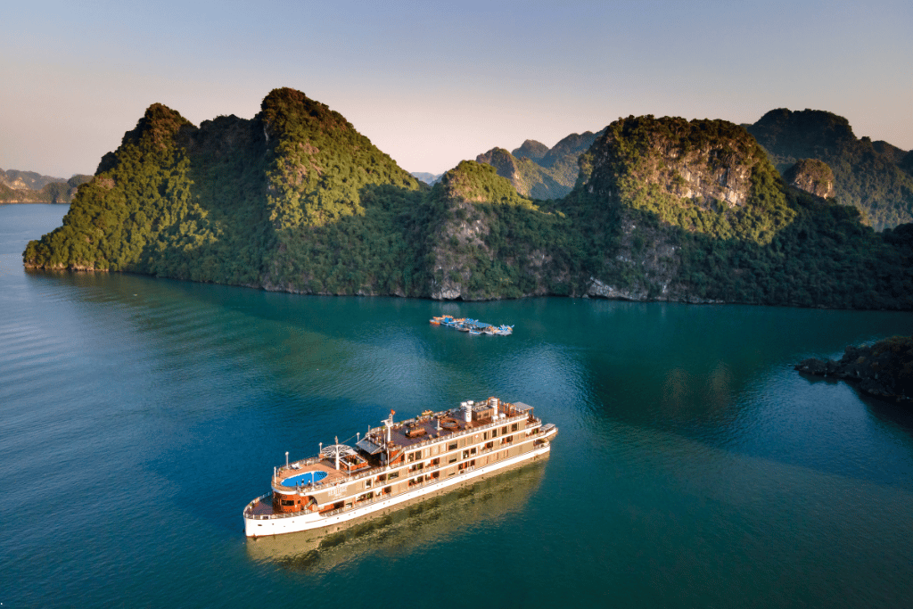 A white multi-deck cruise ship with a rooftop pool moored beside forested limestone islands