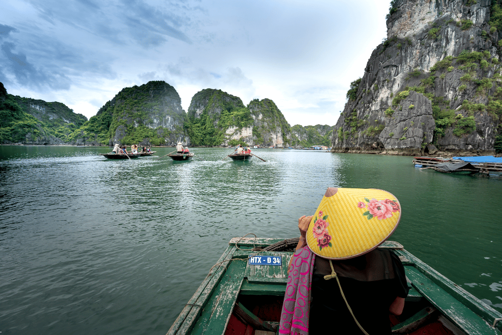 View from a rowed boat as a woman in a yellow conical hat rows towards limestone cliffs