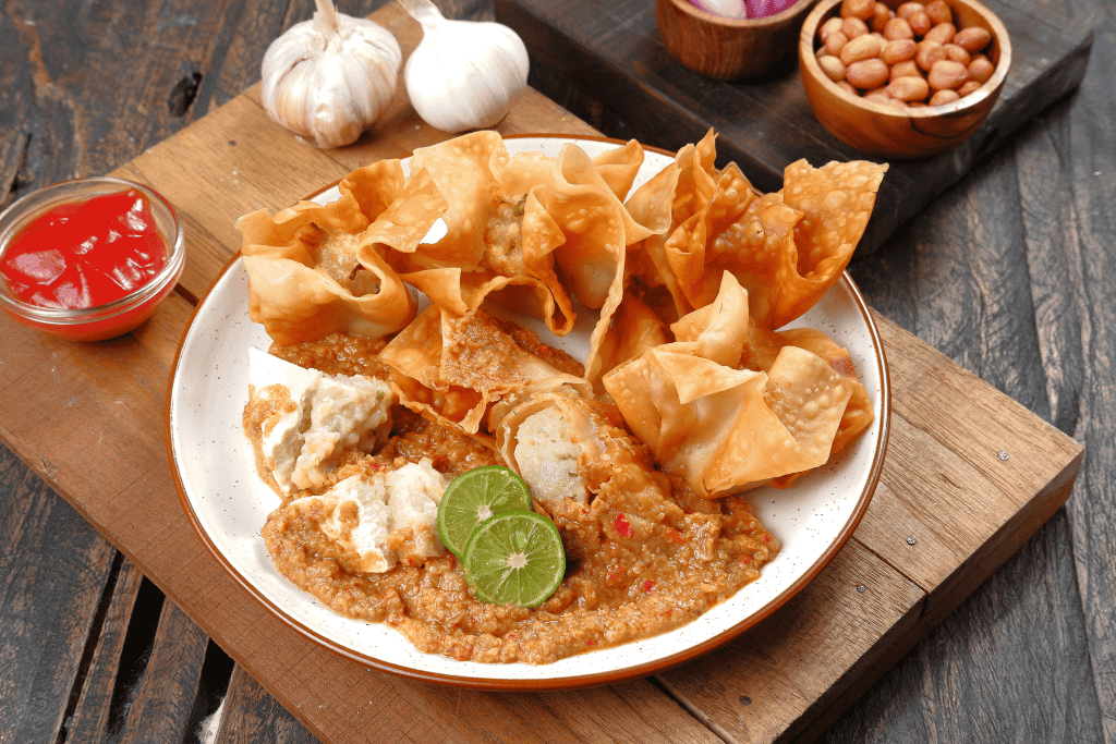 Fried batagor dumplings on a plate under thick peanut sauce with lime halves