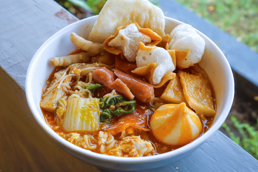 Bowl of seblak in red broth with prawn crackers, sausage, chicken feet and egg