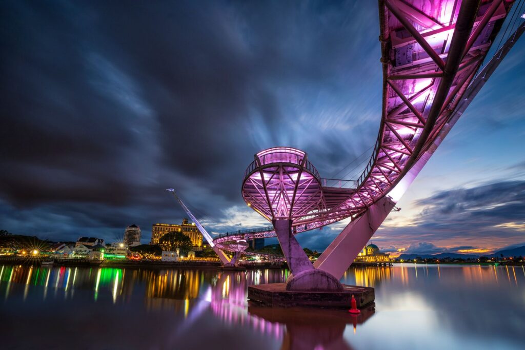Curved steel span of the Darul Hana Bridge lit purple against a deep blue dusk sky