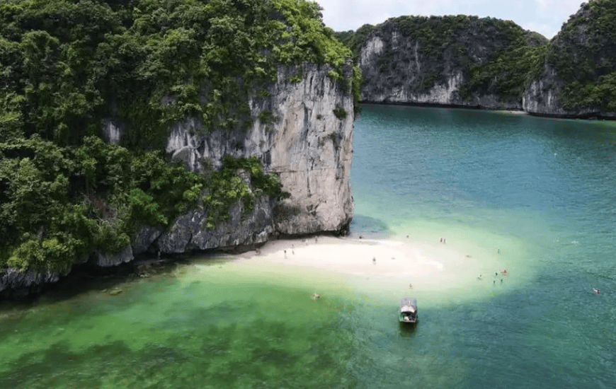 A pale sand spit at the base of a limestone cliff with swimmers and a moored boat