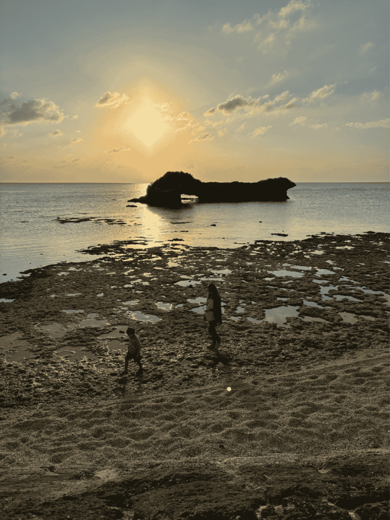 Two children walking across an exposed reef flat at sunset, with an arched rock islet offshore