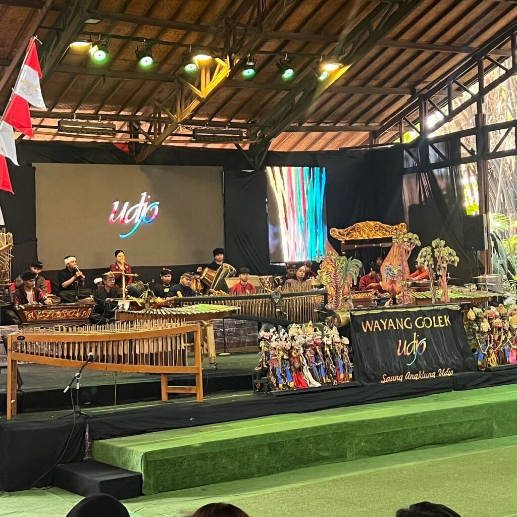 Performers seated behind rows of bamboo angklung instruments on a covered stage