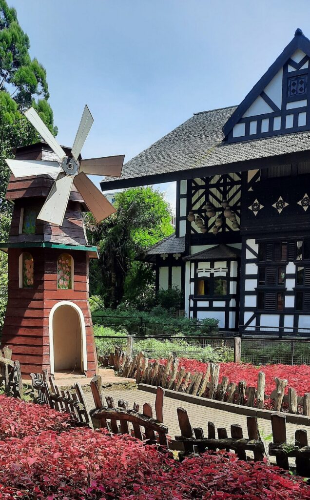 Timbered alpine-style building and a small red windmill above beds of red foliage