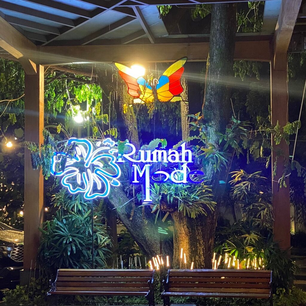 Neon Rumah Mode sign lit at night among trees, benches and a butterfly sculpture