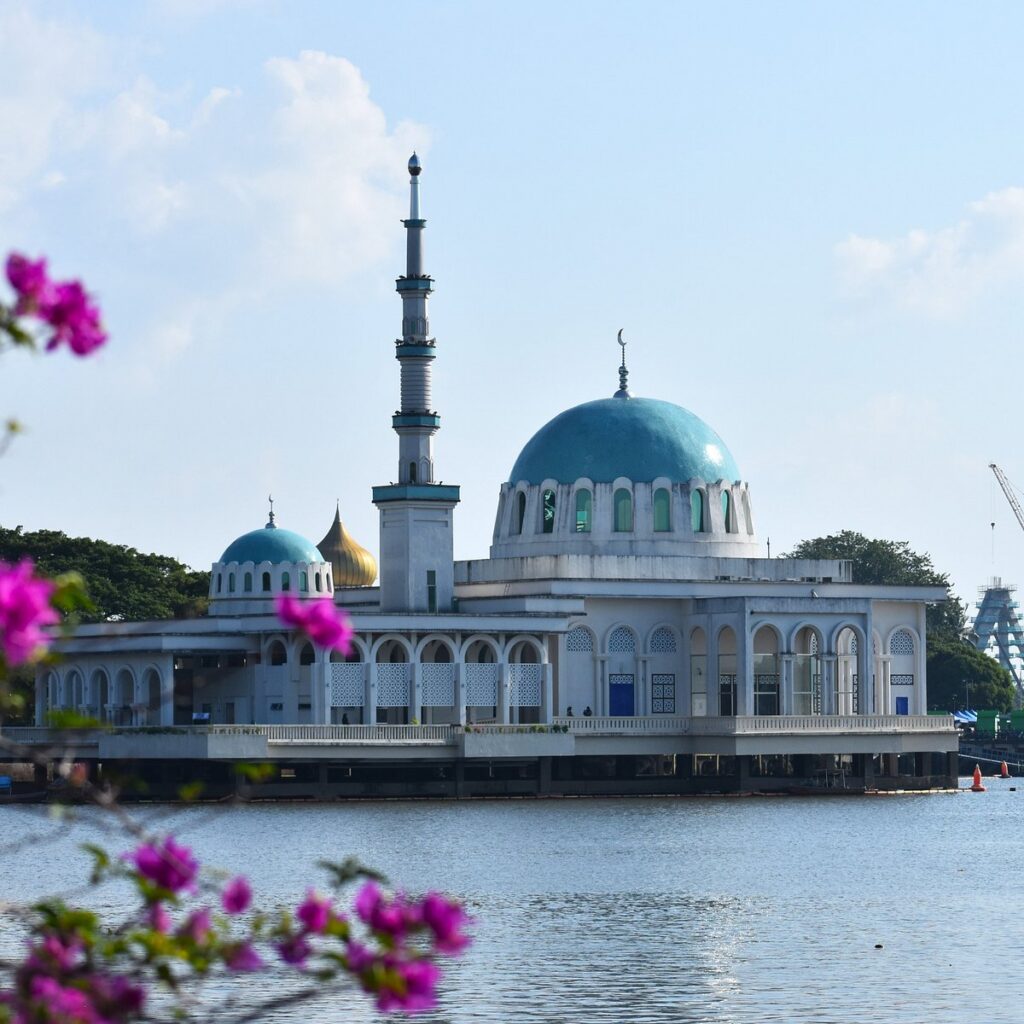 White mosque with turquoise dome and minaret standing over the Sarawak River behind pink bougainvillea