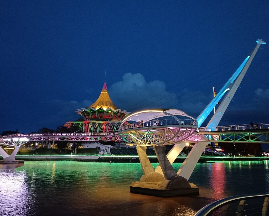 Darul Hana Bridge lit in colour at night with the floodlit State Assembly building behind