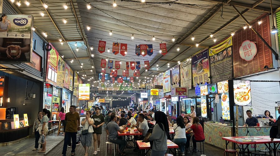 Crowded covered food street lit by string lights, with stalls and shared red tables