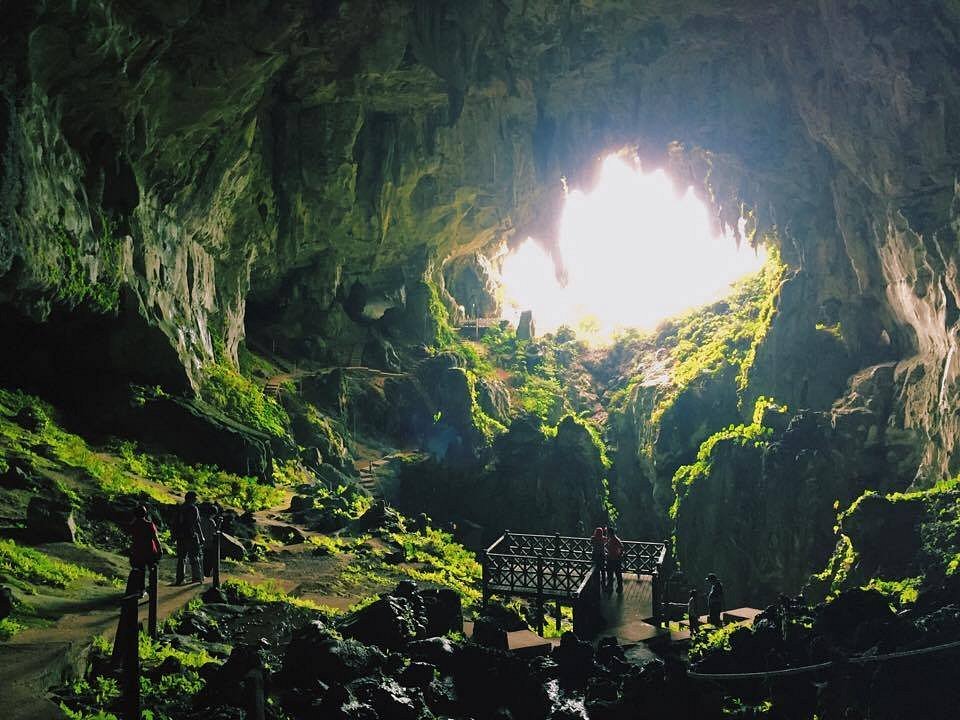Vast cave chamber with mossy rock and daylight flooding through a large opening above a viewing platform