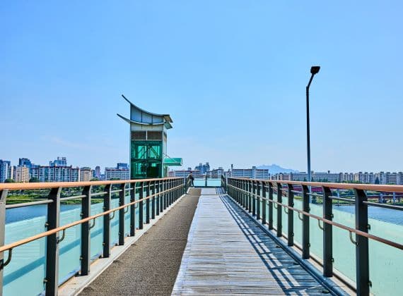 Empty timber-decked footbridge with green railings leading to a small pavilion above a river