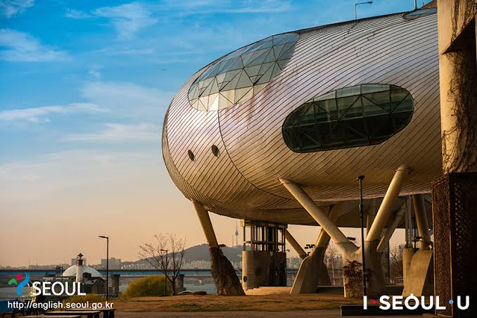 Curved metal-clad building on stilts with oval windows, beside the Han River at sunset
