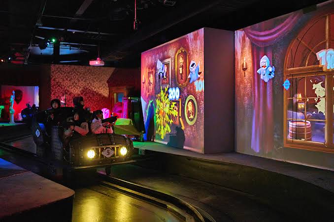 Riders aim light guns from a dark ride car at projected ghosts in a haunted room