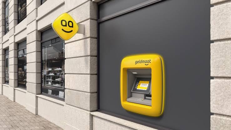 A yellow Geldmaat cash machine set into a dark shopfront wall beside its smiley logo sign