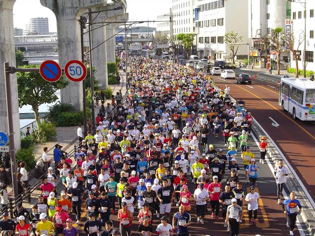 Thousands of runners filling a Naha road beneath the monorail track at the marathon start