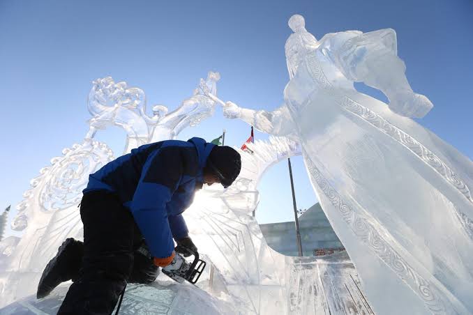 A worker in a blue jacket carving a large ice sculpture with a chisel under blue sky