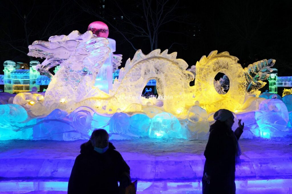 A glowing dragon ice sculpture lit in blue and yellow at night, visitors silhouetted in front