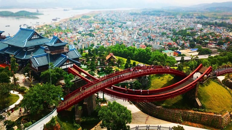 Red curved pedestrian bridge and a blue pagoda on a hillside above a Vietnamese city and bay