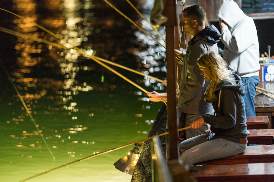 Travellers fishing with rods off a boat deck at night, lamplight reflecting on green water