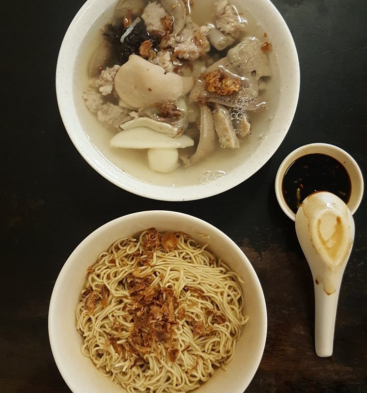 Bowl of dry egg noodles topped with fried shallots beside a clear pork and meatball soup