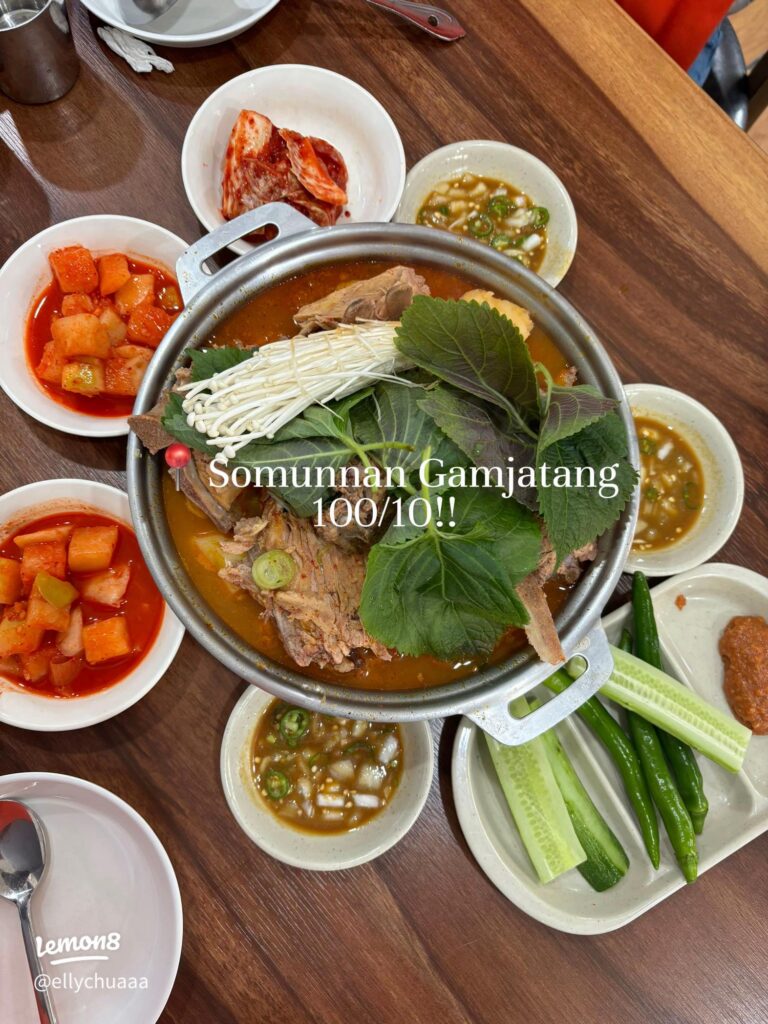 Metal pot of pork bone stew topped with perilla leaves and enoki, ringed by side dishes