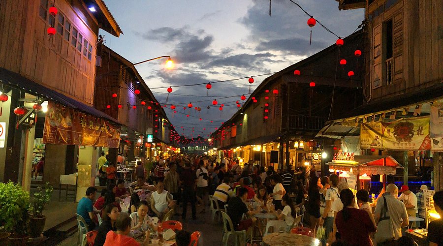 Red lanterns strung above diners at street tables between wooden shophouses at dusk