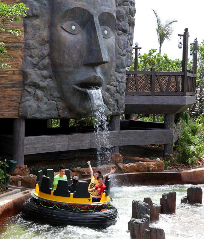 A round raft of riders passing under a carved stone face spouting water on The Rapids ride