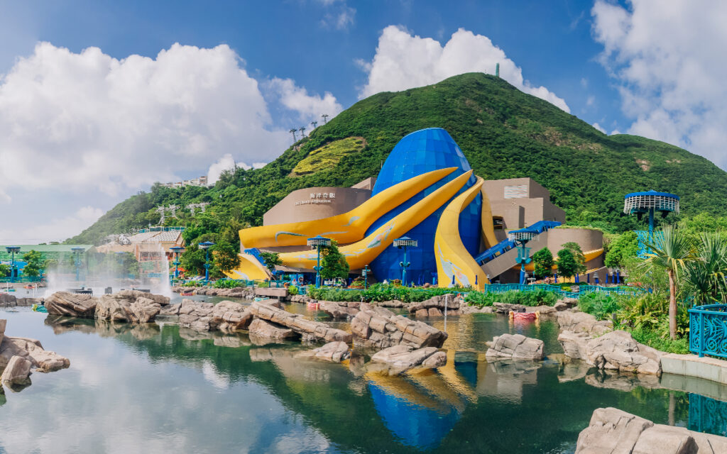 The blue-and-gold Grand Aquarium building reflected in the Aqua City lagoon below the Summit hill