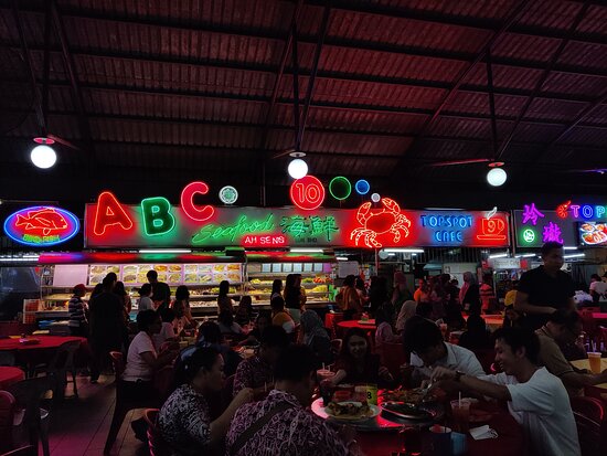 Neon seafood stall signs above busy diners at red tables in an open-air rooftop food court